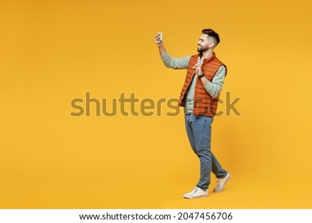 Full length young smiling friendly man in orange vest mint sweatshirt glasses do selfie shot on mobile phone show victory v-sign gesture isolated on yellow background studio. People lifestyle concept