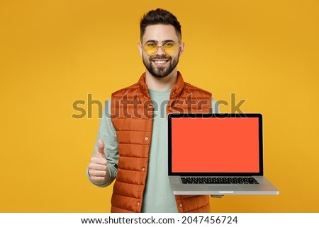 Young freelancer happy man in orange vest mint sweatshirt glasses using laptop pc computer with blank screen workspace area show thumb up like gesture isolated on yellow background studio portrait.
