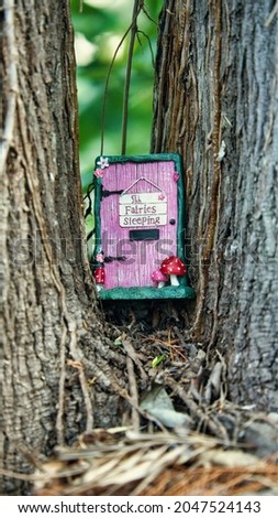 A Fairy House between two trees. A pink and green box showing a door with red mushrooms with white spots including a sign the reads "Shh Fairies Sleeping". The object is positioned centre frame.