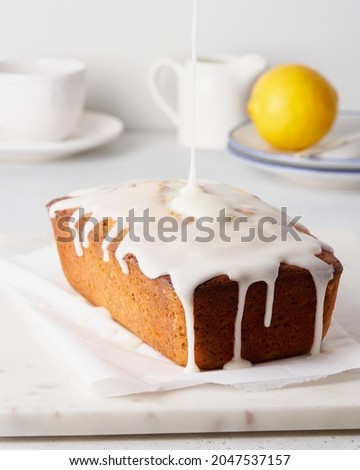 Lemon cake with poppy seeds pouring with white icing. Home made sweet morning breakfast on baking paper and marble tray. Cup of tea and fruit on plate on background. Vertical side view