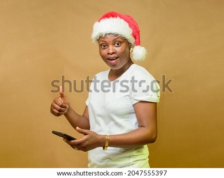 An African woman holding a smart phone, happily looking at the camera, doing thumbs up gesture and also have a Christmas cap on her head