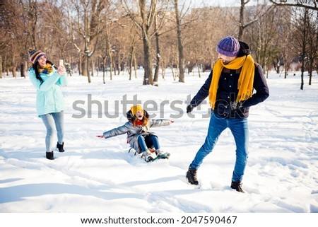 Full size photo of happy cheerful smiling family having good time outdoors little girl riding sledge mother take photo on phone