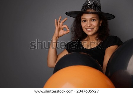 Close-up of beautiful smiling Hispanic woman in wizard hat, dressed in witch carnival costume for Halloween party, shows OK sign, poses against gray background with black balloons with copy space