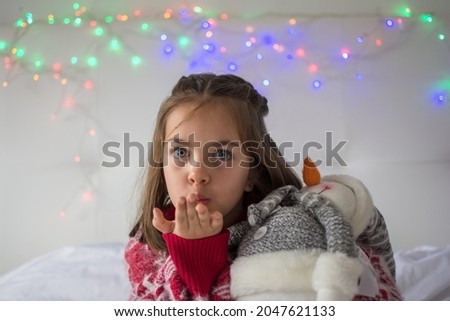 6-Year-Old Girl Blowing A Kiss. Little Girl In Christmas Clothes Hugging A Teddy, With A White Background And Christmas Lights.