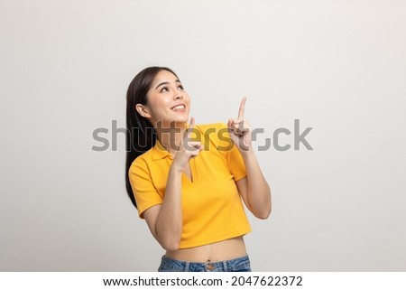 Excited asian woman pointing the finger to blank space over head for advertising text on isolated background. Joyful teenage girl in yellow shirt standing in white room looking at empty space.