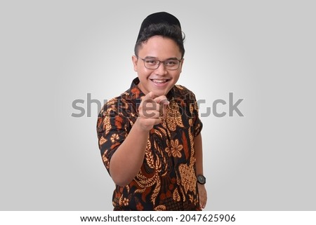 Portrait of smiling Asian man wearing batik shirt and songkok pointing his finger forward to camera. Isolated image on white background