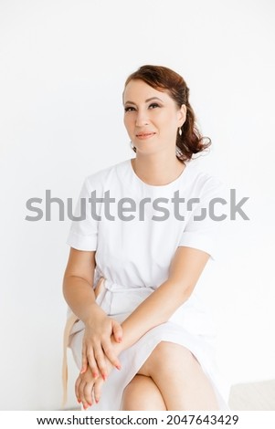 Portrait of a beautiful Caucasian medical girl in a white coat sitting on a stool on a white background. beauty salon.