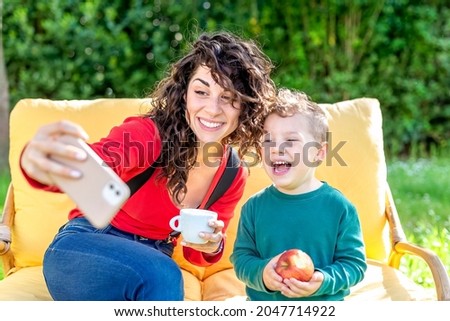 happy mom and son chilling outdoor sitting on a couch in a public park garden making a selfie portrait on smartphone. smiling woman and child interacting online. tech, social media influencer concept