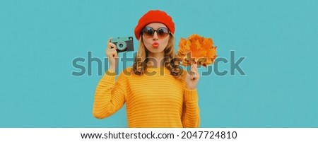 Autumn portrait of beautiful young woman with film camera and yellow maple leaves wearing a sweater, french beret on blue background