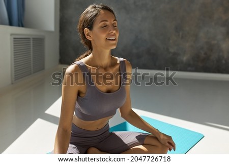 Picture of attractive slim woman in fitness wear sitting in lotus position with eyes closed exhaling with sound showing white healthy teeth, looking happy and relaxed, practicing yoga indoor