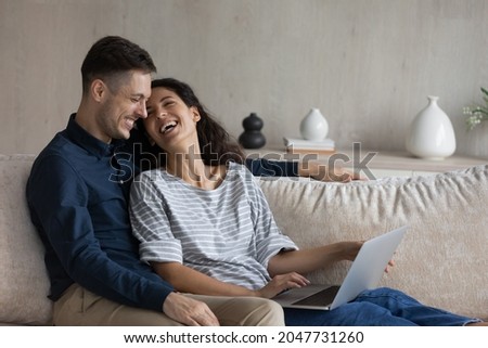 Happy loving young family couple resting using computer software applications, buying goods on services online, playing games or watching funny movie, relaxing together on cozy couch on weekend.