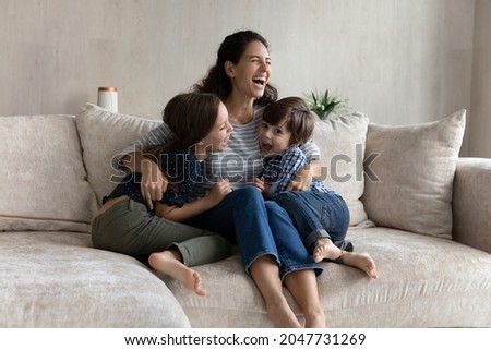 Overjoyed young hispanic mother cuddling small laughing kids siblings, having fun entertaining resting together on comfortable sofa. Joyful multigenerational family playing on weekend at home. Royalty-Free Stock Photo #2047731269