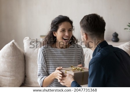 Surprised young hispanic woman taking wrapped box from caring young husband, feeling excited of getting wished present. Joyful millennial man congratulating wife with wedding anniversary or birthday.