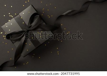 Top view photo of giftbox in black packaging with black ribbon bow and golden sequins on isolated black background with empty space