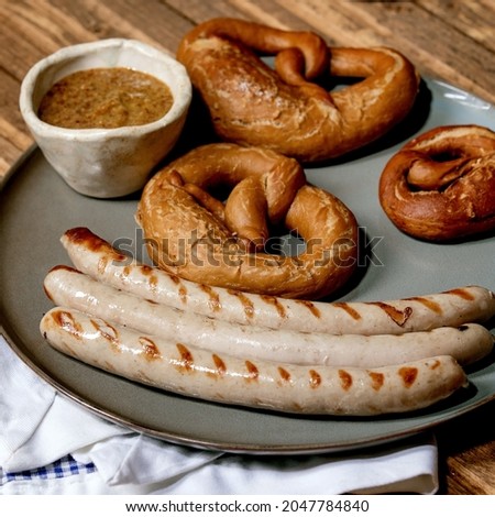 Bavarian traditional grilled pork sausages on ceramic plate served with german sweet mustard, mug of dark beer and pretzels bread on white and blue napkin over wooden background. Square image