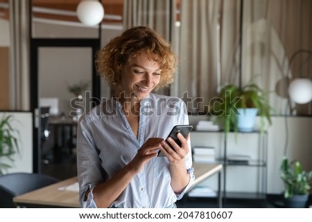 Happy young self employed woman using cellphone alone in office, reading text on screen, making mobile phone call, smiling, chatting online, getting good news, ordering, shopping via online app Royalty-Free Stock Photo #2047810607