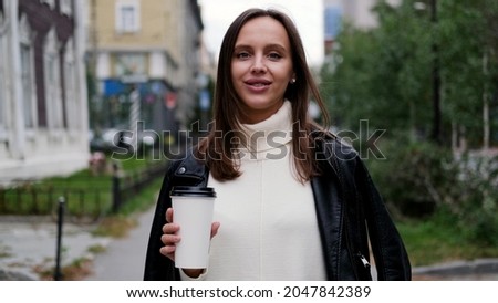 Happy caucasian business woman in a white sweater and black leather jacket walks down the street to her company office holding coffee in her hands
