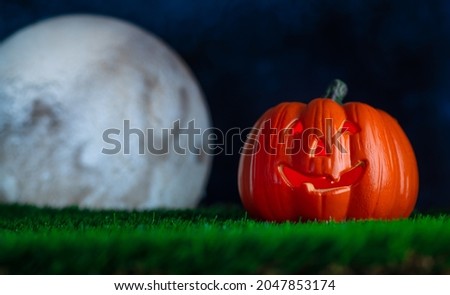 Halloween background. Against the background of a dark night sky and a huge white moon, an orange pumpkin in the form of a smiling head on green grass. Postcard, invitation, banner, advertisement.