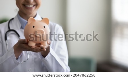Smiling female GP doctor holding piggy bank on hands, showing object at camera, promoting donation, financial insurance, health tax. Saving money, healthcare, finance concept. Cropped shot, close up Royalty-Free Stock Photo #2047862258
