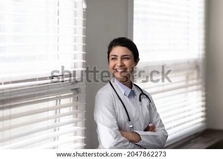 Confident female Indian doctor standing by window, looking away with happy smile. Young general practitioner, therapist thinking of future career vision, enjoying success. Head shot portrait