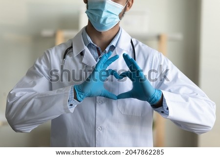 Young male GP doctor in face mask, blue protective latex gloves showing finger heart shape at camera, sending love and support to medical employees and covid patients. Epidemic, medic care concept