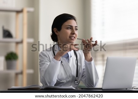 Serious pensive female Indian doctor recording audio message on smartphone, chatting with patient online, giving consultation, advice, explaining recommendation, treatment, using virtual voice app Royalty-Free Stock Photo #2047862420