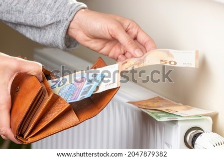 Woman's hand takes euro money banknotes from wallet and places on heating radiator battery with temperature regulator. Expensive heating costs during cold season. Paying bills Royalty-Free Stock Photo #2047879382