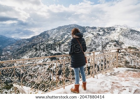 Back view of young woman standing at mountain top observation deck over Vouraikos gorge. Mega Spileon Monastery terrace with splendid snow-covered mountains view. Winter vacation. Greece