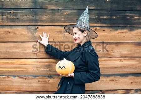 Woman in black witch halloween costume standing with hat and holding a pumpkin isolated on wooden background.