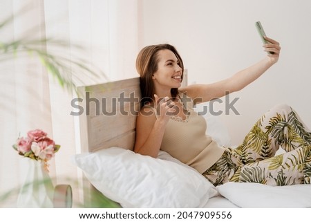 Side view profile young woman in casual casual clothes sit in bed doing selfie shot on mobile cell phone show thumb up like gesture spend time in bedroom lounge home in own room house wake up good day