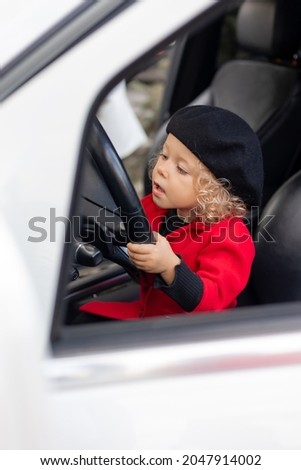 little curly blonde girl with blue eyes in a red coat and a black beret is sitting in the car. child driving parent car. concept for car rental. space for text. High quality photo