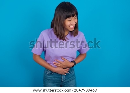 Young beautiful asian girl wearing purple t-shirt over blue background smiling and laughing hard out loud because funny crazy joke with hands on body.