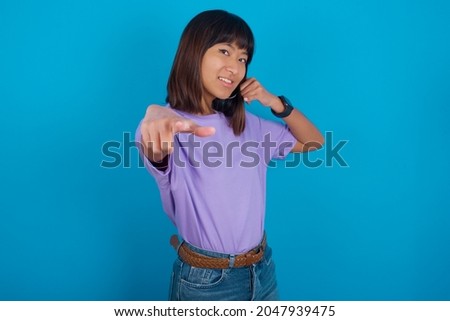 Young beautiful asian girl wearing purple t-shirt over blue background smiling cheerfully and pointing to camera while making a call you later gesture, talking on phone