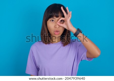 Young beautiful asian girl wearing purple t-shirt over blue background doing ok gesture shocked with surprised face, eye looking through fingers. Unbelieving expression.