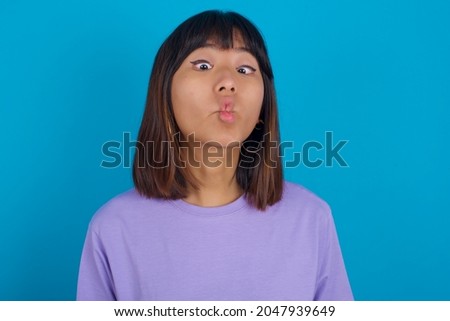 Young beautiful asian girl wearing purple t-shirt over blue background making fish face with lips, crazy and comical gesture. Funny expression.