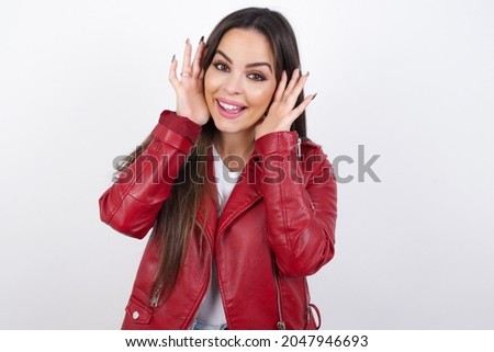 young beautiful brunette woman wearing red biker jacket over white wall Pleasant looking cheerful, Happy reaction