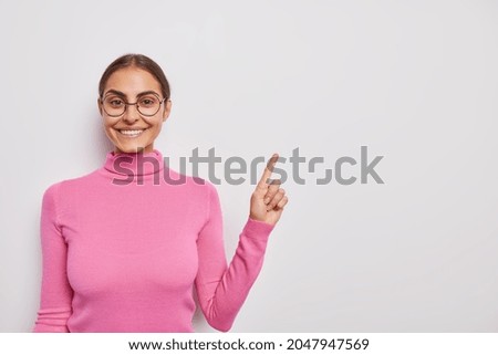 Positive dark haired woman with cheerful expression says look here indicates at upper right corner advertises your text poses over white background wears pink turtleneck and round spectacles