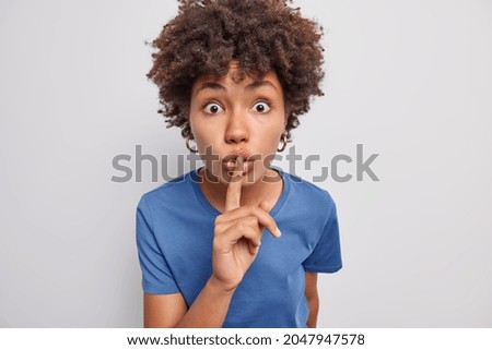 Surprised African American woman with curly hair makes silence gesture keeps finger pressed to lips shows hush sign dressed in casual blue t shirt isolated over white background. Shush be quiet