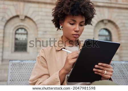 Creative Afro American woman works on tablet with stylus draws sketches for future project works on editing pictures for image productionn sits outdoors wears stylish clothes. Female web designer