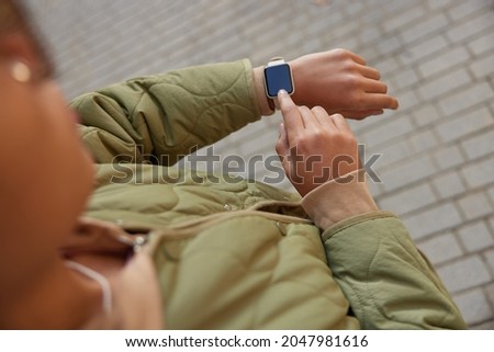 Cropped shot of unrecognizable stylish woman checks notification on digital wearable wrist watch monitors time wears casual jacket poses at street. Faceless female uses modern gadget at street Royalty-Free Stock Photo #2047981616