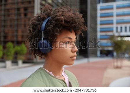 Sideways shot of curly haired woman in wireless headphones listens audio track while walking in city poses against blurred background enjoys spare time thinks about plans on weekend. Lifestyle