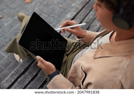 View from back of unrecognizable young woman sits on wooden bench uses touch pad and application for editing photos holds tablet with blank empty screen white stylus takes notes poses outside