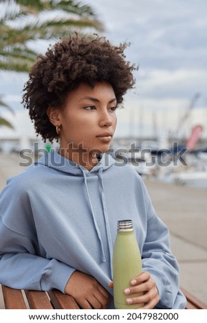 Vertical shot of thoughtful woman rests after workout feels thirsty holds bottle of water dressed in hoodie poses outdoors against harbor background enjoys rest during spare time. Healthy lifestyle