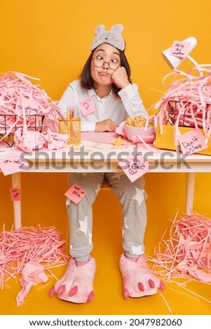 Funny Asian woman makes grimace crosses eyes sits at desktop dressed in pajama works from home poses against yellow background. Female student prepares for exam studies at domestic atmosphere