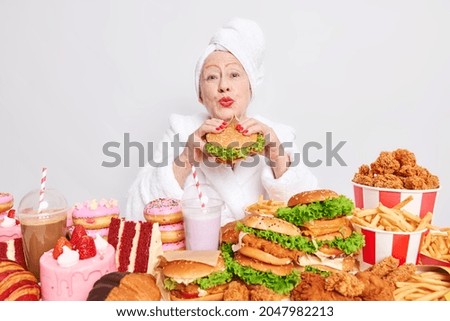 Wrinkled elderly woman keeps lips rounded wants to kiss you eats delicious hamburger has fast food addiction dressed in bathrobe isolated over white background. High calorie snacks and cheat meal