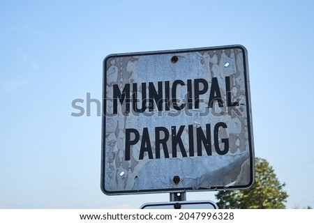 An old metal sign is labeled 'Municipal Parking' in large black lettering and seem from a low angle against sky. The sign shows damage from the weather and sunshine, forming varied textures across it.