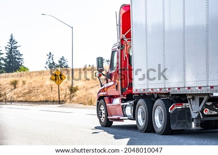 Day cab model red big rig semi truck with aerodynamic roof spoiler transporting commercial cargo in dry van semi trailer running on the summer highway road with road sign and yellow hill Royalty-Free Stock Photo #2048010047