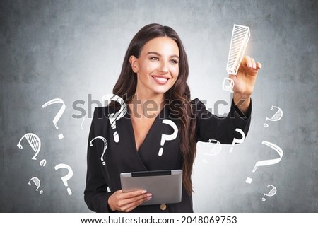 Smiling businesswoman wearing formal suit is touching exclamation mark with her finger. Sketch with lots of question marks on concrete wall in background. Concept of searching for solution to problem