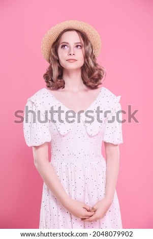 A pretty girl in a light white summer dress and a straw hat looking up dreamily in a pink background. Summer beauty and fashion. Happy people.
