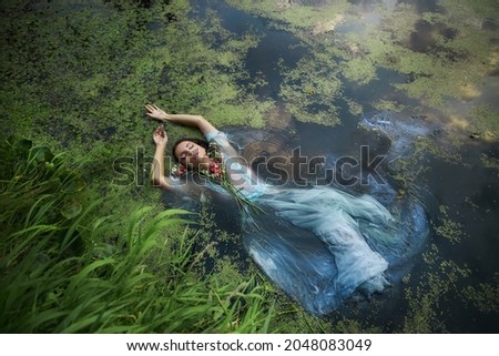 Art beautiful romantic woman lies in swamp in blue long dress with flowers. Portrait brunette in transparent dress in water swamp mud duckweed. Book cover Royalty-Free Stock Photo #2048083049
